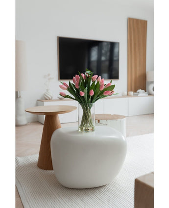 Een moderne woonkamer met een wit vloerkleed, een grote ovale salontafel en een kleine ronde houten bijzettafel. Op de tafel staat het Zijden kunstboeket Tulpen in vaas Roze; een tv en een minimalistisch decor maken de achtergrond compleet.