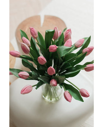 Het Zijden kunstboeket Tulpen in vaas Roze toont roze kunsttulpen met groene bladeren, gerangschikt in een glazen vaas, tentoongesteld op een wit vlak met een zacht vervaagde lichte achtergrond, van bovenaf gezien.