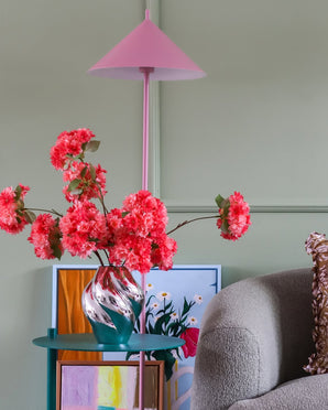 Une lampe rose, un vase rayé avec le bouquet de fleurs artificielles en soie, des fleurs de cerisier et du corail, des œuvres d'art colorées et une chaise grise se dressent contre un mur vert clair, créant un coin confortable et animé.