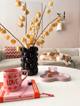 Un salon cosy avec un canapé beige, un chat endormi, un vase noir sculptural contenant la Fleur Artificielle Physalis Lampion 90 cm marron, un mug rose "LOVE", un livre, une bougie et un biscuit sur la table basse.