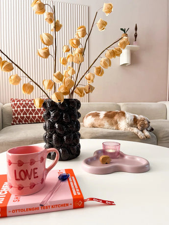 Un salon cosy avec un canapé beige, un chat endormi, un vase noir sculptural contenant la Fleur Artificielle Physalis Lampion 90 cm marron, un mug rose "LOVE", un livre, une bougie et un biscuit sur la table basse.